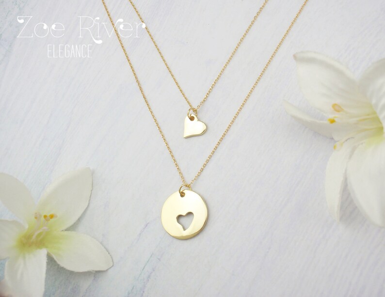 May include: Two gold-colored necklaces. The top necklace has a small heart pendant. The bottom necklace has a circular pendant with a heart-shaped cutout. The necklaces are layered and displayed with white flowers. The text "Zoe River Elegance" is in the upper left corner.