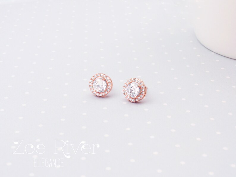 May include: A pair of rose gold stud earrings with a halo of small, clear crystals surrounding a larger, central crystal. The earrings are displayed on a light gray surface with white polka dots. The brand name "Zoe River Elegance" is visible.