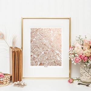 May include: A framed print of a rose gold map of Paris. The map is surrounded by a white mat and a gold frame. The print is displayed on a white surface with a bouquet of pink roses, a stack of books, and a pearl necklace.