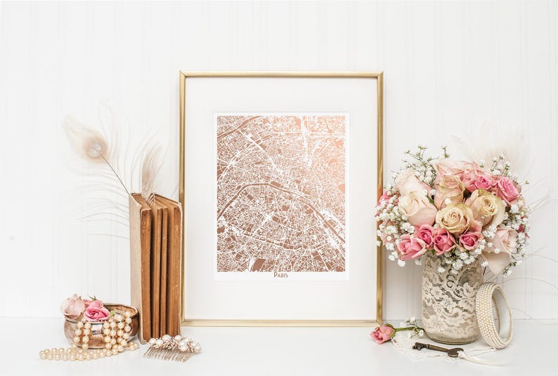 May include: A framed print of a rose gold map of Paris. The map is surrounded by a white mat and a gold frame. The print is displayed on a white surface with a bouquet of pink roses, a stack of books, and a pearl necklace.