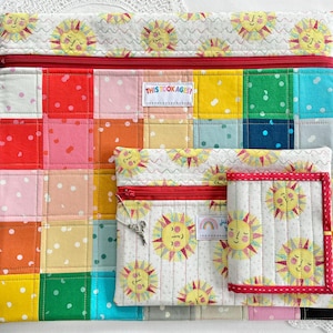May include: A collection of quilted fabric pouches in different sizes. The largest pouch has a patchwork design with red, pink, orange, yellow, green, and blue squares. Smaller pouches feature smiling suns on a white background. All have red zips.
