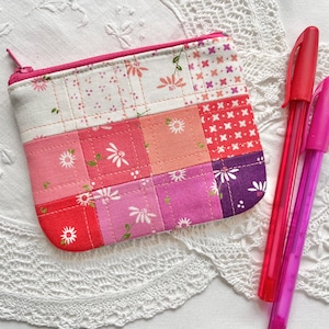 May include: A patchwork coin purse with a pink zip, displaying floral and geometric patterns in pink, red, and purple hues. Two pens, one red and one pink, are placed beside the purse.
