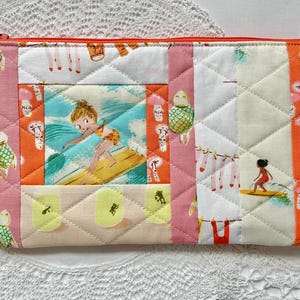 May include: Quilted fabric pouch with a zip closure. The patchwork design features a surfer girl, turtles, and other tropical motifs in shades of pink, orange, and white. The pouch is quilted with a diamond pattern.