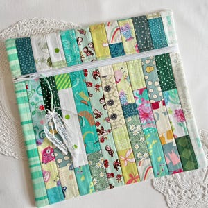 May include: A square zippered pouch constructed from vertical fabric strips in various shades of green, teal, and white. The fabric features diverse patterns including owls, flowers, and geometric shapes. A white lace ribbon is tied to the zipper pull.