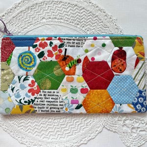 May include: A rectangular zipped pouch constructed from quilted fabric hexagons in various colours and patterns, including floral, polka dot, and fruit prints. A small purple zip pull is attached to the top left corner of the pouch.
