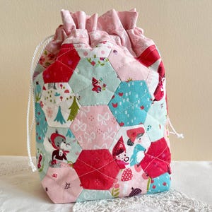 May include: A patchwork drawstring bag in shades of red, pink, and teal. The bag is made from hexagonal fabric pieces with various patterns and illustrations. It has a white drawstring closure and a gathered top.