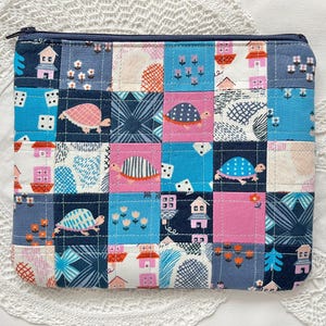 May include: A patchwork pouch with a quilted design. The design features turtles, houses, and floral patterns in blue, pink, and white. It has a navy blue zip. The pouch is likely used for storing small items.