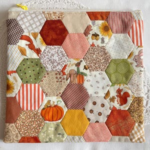 May include: A quilted pouch with a patchwork design featuring autumn-themed patterns. The hexagonal patches display squirrels, pumpkins, sunflowers, and leaves in shades of orange, brown, and green. A yellow zip secures the top.