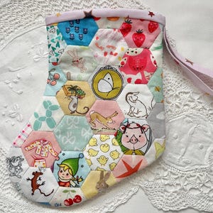 May include: A handmade patchwork stocking with a variety of colourful hexagonal patches. The patches feature whimsical illustrations of animals, strawberries, and other playful designs. A lavender strap is attached to the top.