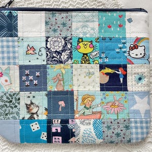 May include: A patchwork zipped pouch featuring a variety of blue and white patterned squares. The squares display whimsical illustrations of cats, bunnies, and other playful designs. The pouch has a dark blue zip.