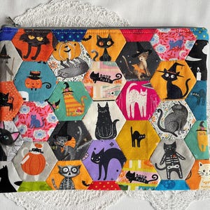 May include: A rectangular zipped pouch featuring a patchwork design of colourful fabric hexagons. Each hexagon displays various illustrations of cats, some wearing witch hats or costumes, along with pumpkins and the text Sour Puss.