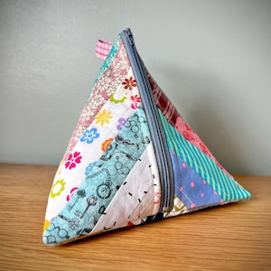 May include: A handmade, triangular fabric pouch with a grey zip. The pouch is made from various patterned fabric scraps, including floral, key, and polka dot designs. The pouch is resting on a wooden surface.