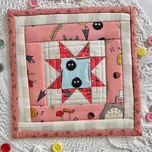 May include: A square, handmade quilted coaster with a star design, a light blue centre, and two black, fluffy characters. The coaster has a pink background with cartoon illustrations and a white and pink checkered border. Colourful buttons surround the coaster.