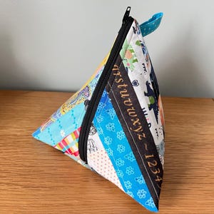 May include: A triangular fabric pouch featuring a patchwork design with various colourful patterns, including a black strip printed with the alphabet letters a through z and numbers 1 through 4. It has a black zip closure and a small blue loop.