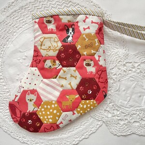 May include: A handmade Christmas stocking with a patchwork design. It features dog illustrations on red, white, and gold hexagonal fabric pieces. The stocking has a striped loop for hanging.