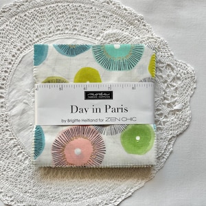 May include: A stack of square fabric pieces wrapped in a paper band labelled Day in Paris by Brigitte Heitland for ZEN CHIC. The fabric features a white background with circular patterns in teal, lime green, and pink. A ruler shows a width of 12.7 cm.