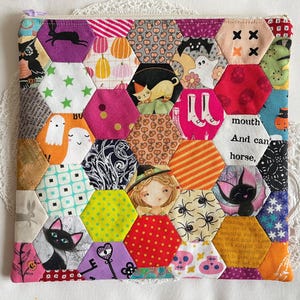 May include: A patchwork zipped pouch with a Halloween theme. The hexagonal fabric pieces feature spooky images such as ghosts, cats, and witches. The text "mouth And can' horse," is visible. The pouch has a purple zip.
