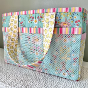 Könnte beinhalten: Eine rechteckige Stofftasche mit hellblauem Hintergrund und einem Blumen- sowie Hasenmuster. Die Tasche verfügt über zwei Tragegriffe mit gelbem Kükenmuster, eine hellblaue Netztasche auf der Vorderseite und eine gestreifte Einfassung.
