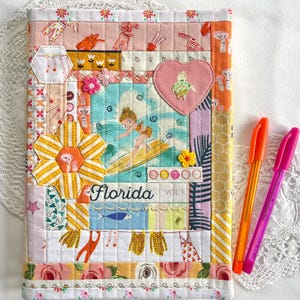 May include: A patchwork fabric journal cover with a surfer girl illustration and the word "Florida". The cover includes a heart with a turtle design. Two pens, one orange and one pink, are placed beside the journal.