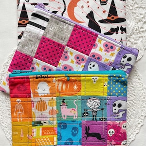 May include: Three colourful Halloween-themed zipped pouches. The top pouch is white with black witch hats, a black cat, and an orange crescent moon. The middle pouch has pink and grey squares. The bottom pouch has a patchwork design with Halloween characters.