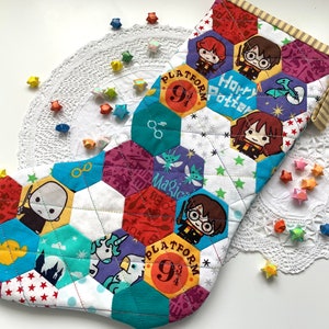 May include: A colourful, handmade Christmas stocking with a patchwork design. The stocking features hexagonal patches with Harry Potter characters and symbols. The words "Platform 9 3/4" are visible. The stocking is decorated with star-shaped origami and rests on a white doily.