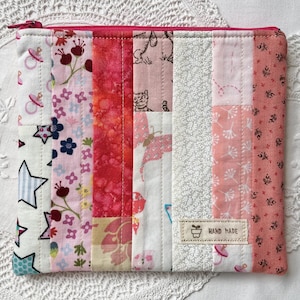 May include: A rectangular zipped pouch constructed from vertical fabric strips in various shades of pink, white, and grey. The fabric features patterns including stars, flowers, butterflies, and pacifiers. A small beige label reads HAND MADE.