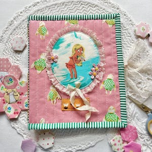 May include: A pink fabric journal cover with a beach scene illustration, green striped trim, and a decorative bow. The cover features a central oval design with a girl in the water, surrounded by a lace border and floral accents. Additional fabric embellishments are scattered around.