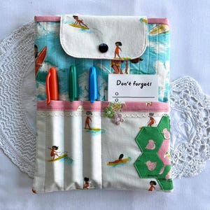 Puede incluir: Organizador de tela hecho a mano con temática de surf. Cuenta con una solapa con botón, un bolsillo para bolígrafos y un bloc de notas con "Don't forget!". El diseño incluye surfistas y olas.