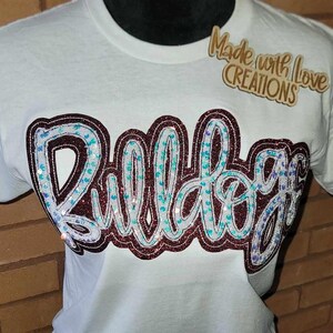 May include: White t-shirt featuring a maroon and white sequined "Bulldog" design. The shirt also has a small wooden tag that reads "Made with Love Creations".