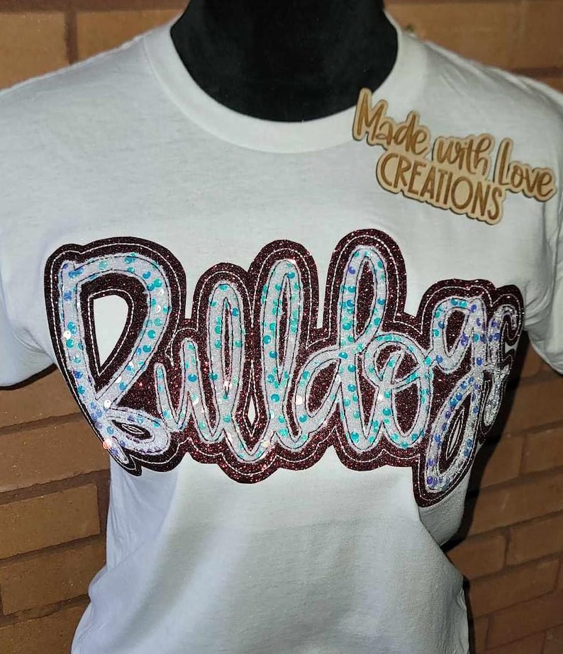 May include: White t-shirt featuring a maroon and white sequined "Bulldog" design. The shirt also has a small wooden tag that reads "Made with Love Creations".