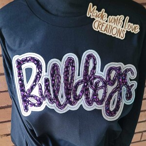 May include: Black long-sleeved shirt featuring a purple sequined "Bulldogs" design. The shirt has a white outline around the design. The text "Made with Love Creations" is visible in the top right corner of the image.