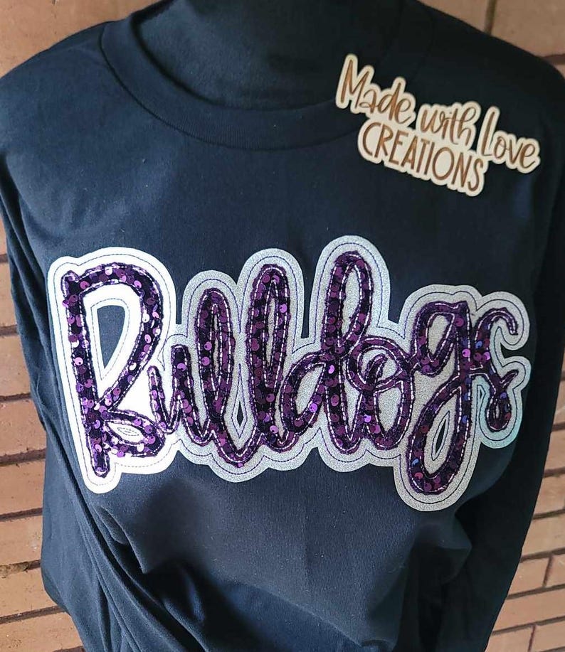 May include: Black long-sleeved shirt featuring a purple sequined "Bulldogs" design. The shirt has a white outline around the design. The text "Made with Love Creations" is visible in the top right corner of the image.