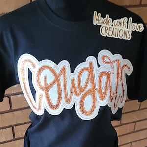 May include: Black t-shirt with a copper glitter iron-on design that says "Cougar" with the words "Made with Love Creations" in the top right corner.