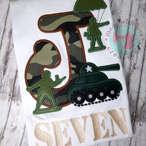 May include: A white t-shirt with a green camouflage letter J, a green toy soldier parachuting, a green toy soldier standing, and a green toy tank. The text "SEVEN" is embroidered in gold below the design. The shirt is decorated with the text "made with love creations".