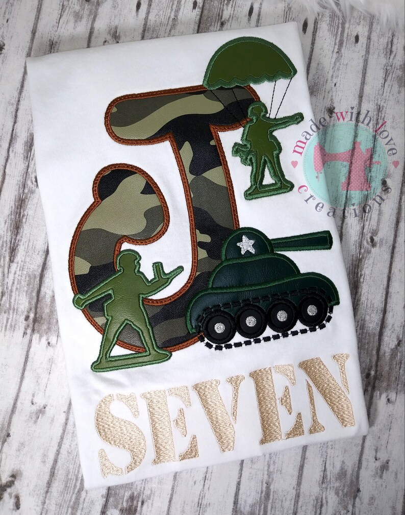 May include: A white t-shirt with a green camouflage letter J, a green toy soldier parachuting, a green toy soldier standing, and a green toy tank. The text "SEVEN" is embroidered in gold below the design. The shirt is decorated with the text "made with love creations".