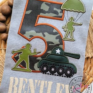 May include: A gray t-shirt with a camouflage number 5, a green tank, a soldier with a parachute, and a soldier holding a gun. The text "BENTLEY" is embroidered below the design. The shirt has a circular stamp that says "Made with Love".