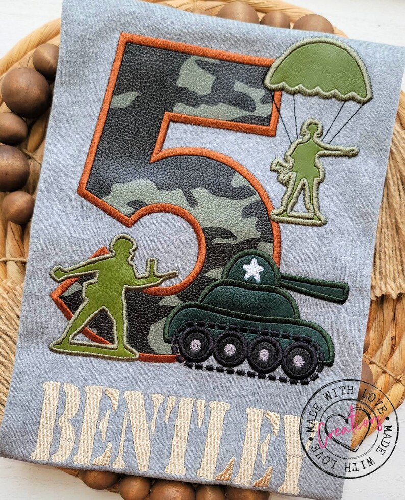 May include: A gray t-shirt with a camouflage number 5, a green tank, a soldier with a parachute, and a soldier holding a gun. The text "BENTLEY" is embroidered below the design. The shirt has a circular stamp that says "Made with Love".