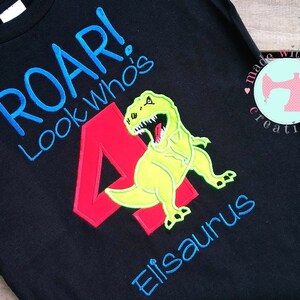 May include: Black t-shirt with a green dinosaur and a red number 4. The text "Roar! Look who's" is embroidered in blue on the shirt. The text "Elisaurus" is embroidered in blue below the dinosaur.