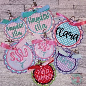 May include: Seven colorful round keychains with scalloped edges. Each keychain features a different name or monogram, including Hayden Ella, Clara, SSU, Harper, and Mila James. The keychains are decorated with gingham, polka dots, and glitter.