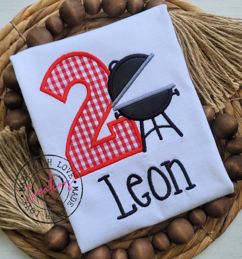 May include: A white t-shirt with a red and white checkered number two and a black grill with a gray lid. The name Leon is embroidered below the grill. The shirt has a small pink heart with the text "Made with Love" on it.