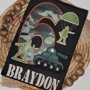 May include: A black t-shirt with a camouflage number six, a soldier with a parachute, a soldier with a gun, a tank, and the name Braydon in white text.