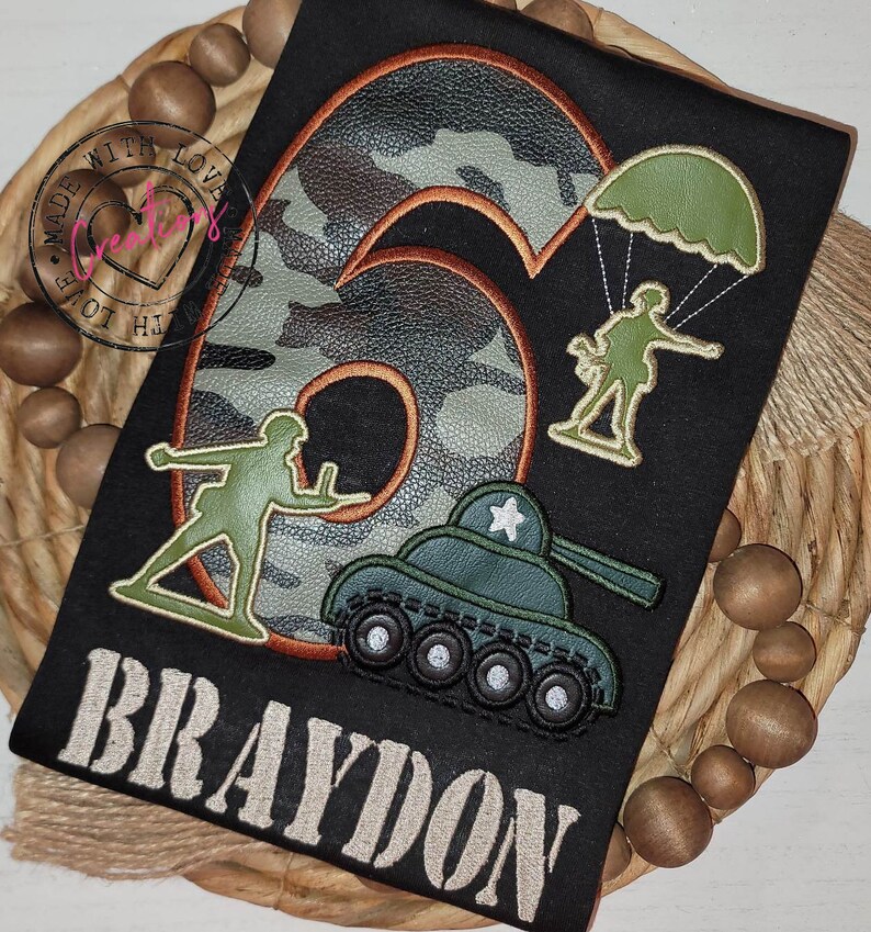 May include: A black t-shirt with a camouflage number six, a soldier with a parachute, a soldier with a gun, a tank, and the name Braydon in white text.