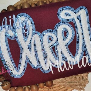 May include: A maroon shirt with white and blue sequined lettering that reads "Unity Cheer Mamaw".