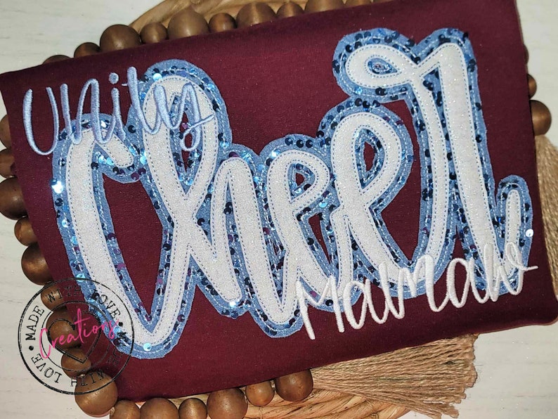 May include: A maroon shirt with white and blue sequined lettering that reads "Unity Cheer Mamaw".