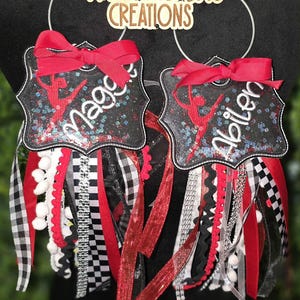 May include: Two custom-made homecoming mums with red bows, personalized names, and a dancer silhouette. The mums feature black and white checkered ribbons, red ribbons, and white pom-poms. The top reads "Made with Love CREATIONS".
