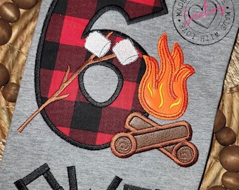 May include: A gray t-shirt with a red and black plaid number six, a campfire with flames and logs, and three white marshmallows on a stick. The text "Owen" is below the number and the words "Made with Love" are in a circle on the right side of the shirt.