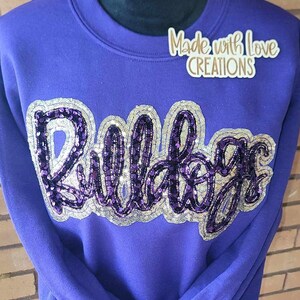 May include: Purple sweatshirt with the word "Bulldogs" in silver and purple sequins. The sweatshirt has a crew neck and long sleeves. The text "Made with Love Creations" is on the top right corner of the sweatshirt.