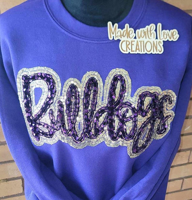 May include: Purple sweatshirt with the word "Bulldogs" in silver and purple sequins. The sweatshirt has a crew neck and long sleeves. The text "Made with Love Creations" is on the top right corner of the sweatshirt.