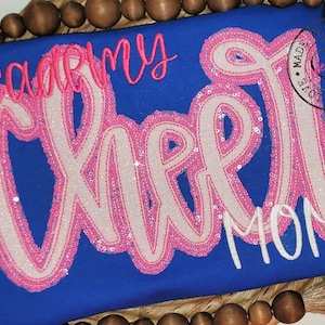 May include: A royal blue t-shirt with the words "Academy Cheer Mom" in pink and white sequins. The word "Cheer" is larger and in a cursive font.