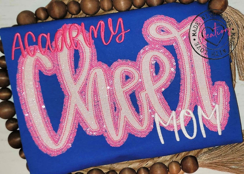 May include: A royal blue t-shirt with the words "Academy Cheer Mom" in pink and white sequins. The word "Cheer" is larger and in a cursive font.